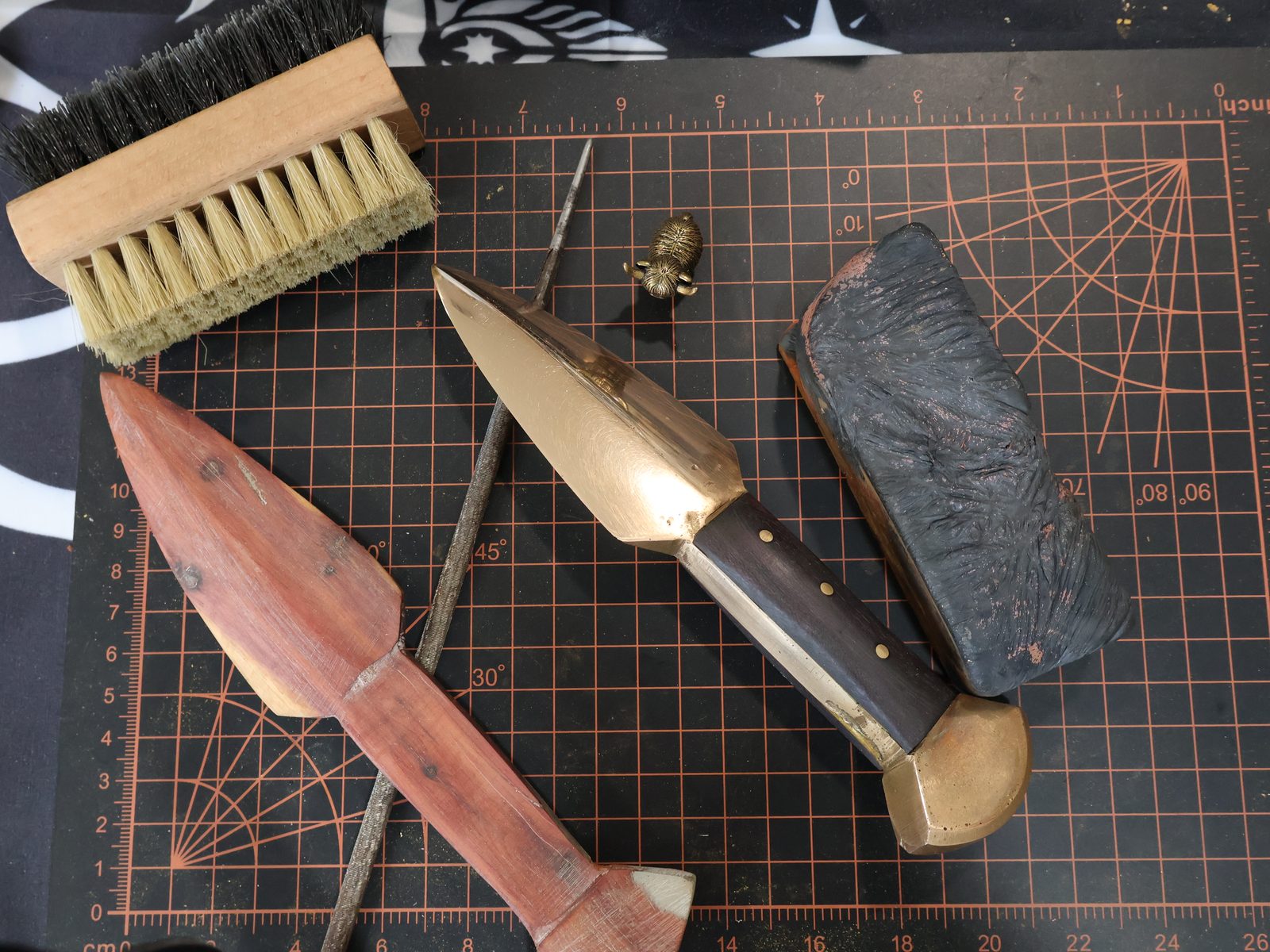 The bench from above: wire brush, files, a carved wooden pattern and a finished bronze knife with a dark grip laid across a cutting mat