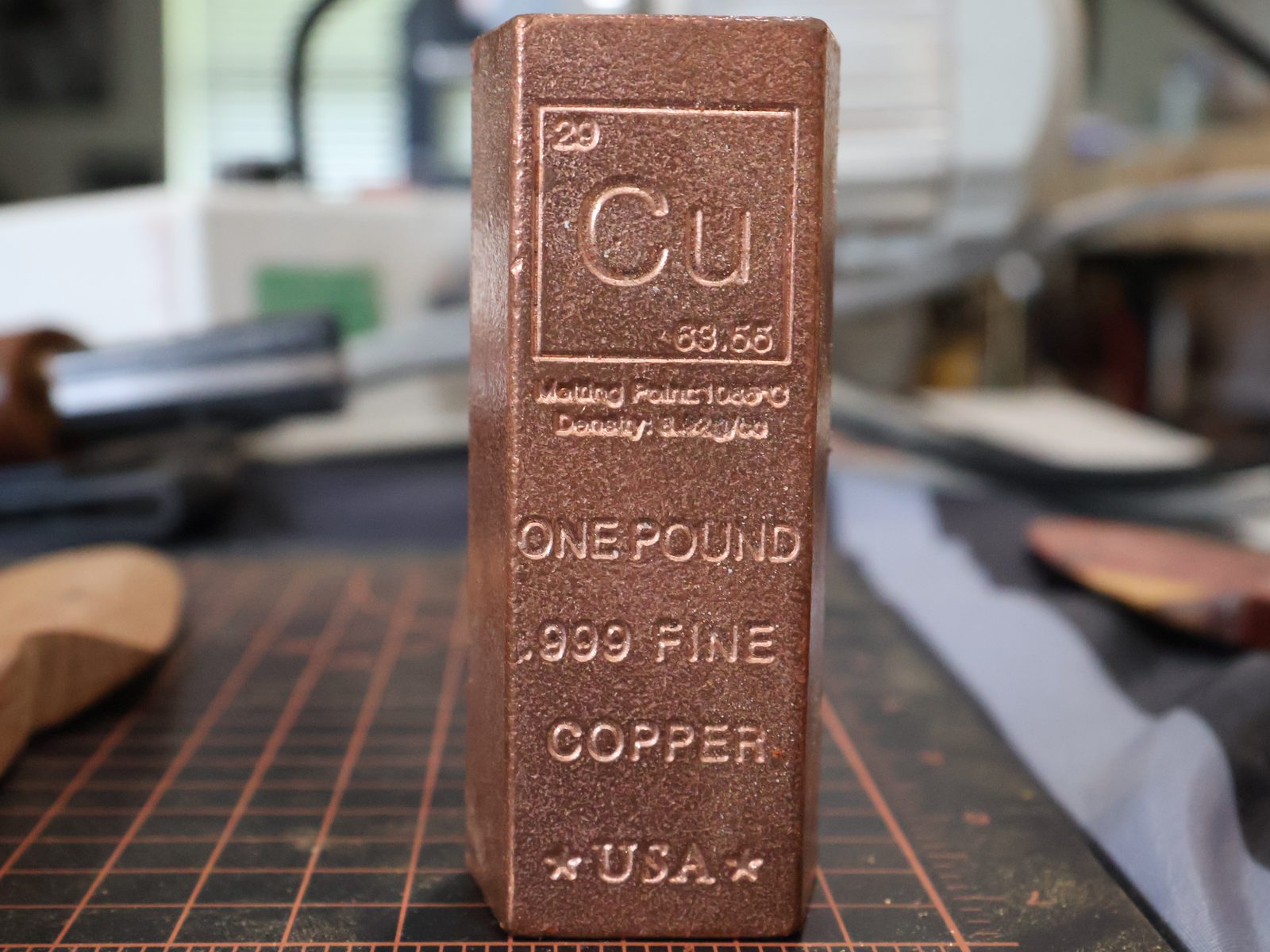 A one-pound bar of .999 fine copper stamped with the element symbol Cu, standing on a cutting mat at the bench