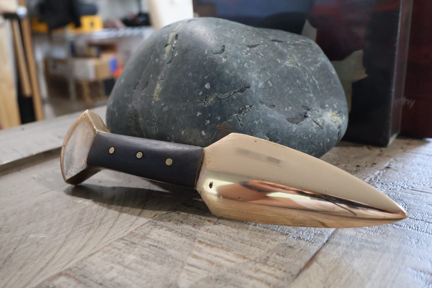 The knife propped against a grey river stone, showing the swell of the blade and the flared bronze pommel