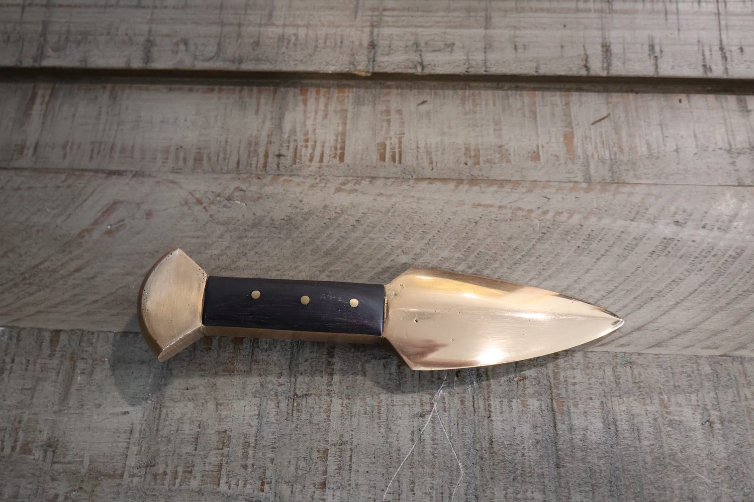 The knife from directly above on weathered timber: leaf-shaped bronze blade, dark grip with three brass pins, flared pommel
