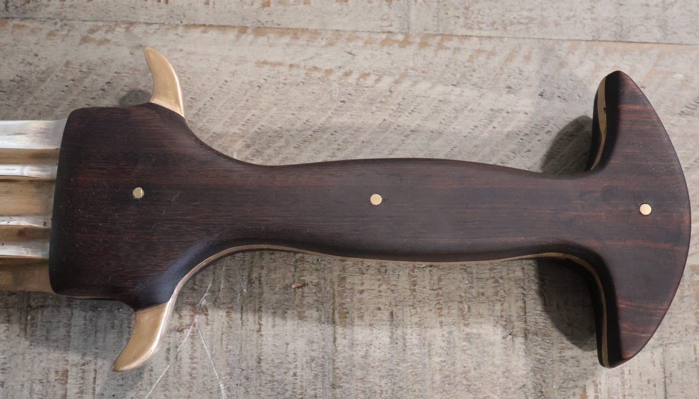 Top-down view of the sword's dark hardwood grip, three brass pins down its length, a cast bronze quillon curling out at the guard and a wide flared pommel at the end