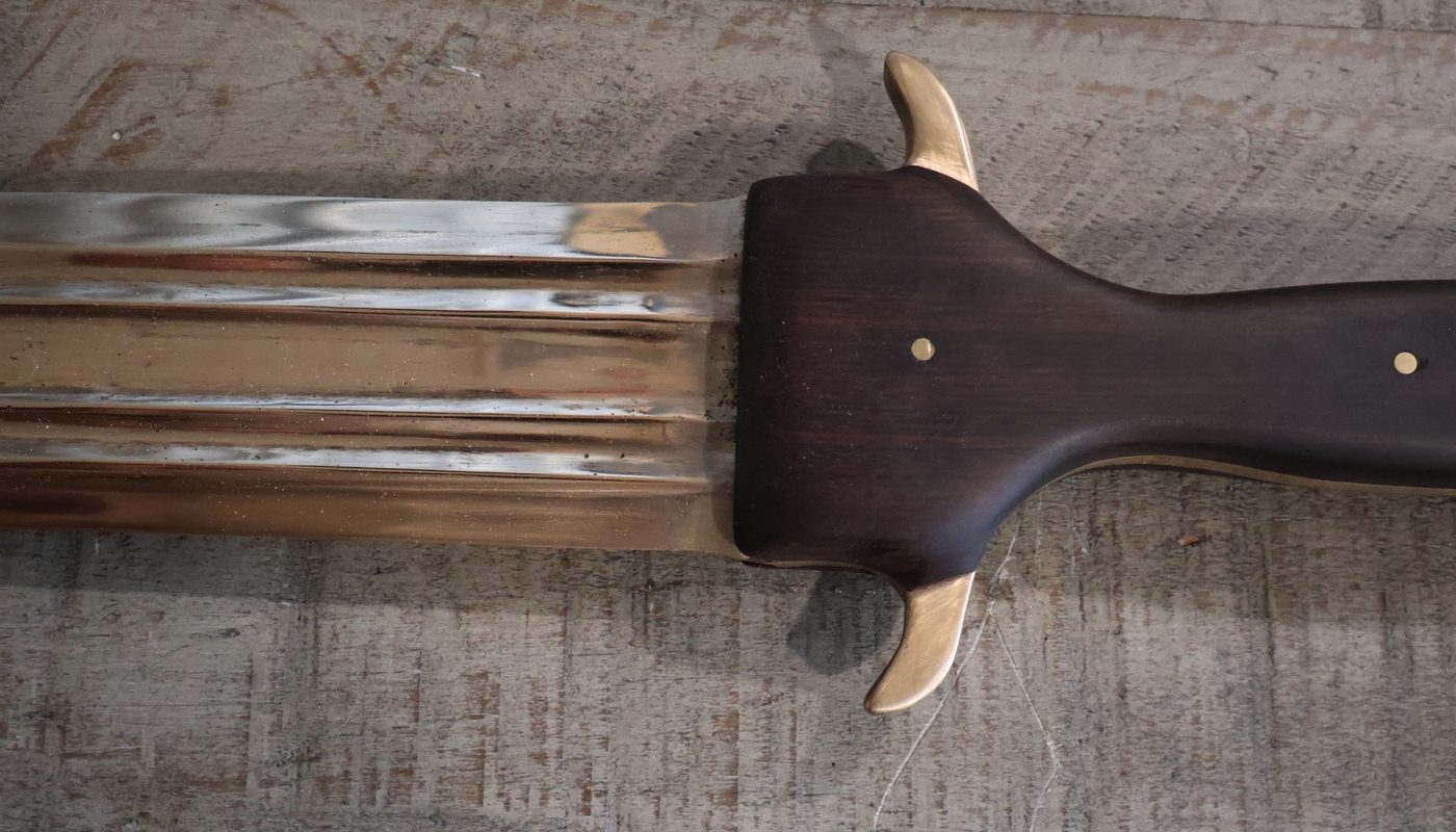 The guard where blade meets handle: polished bronze fullers running out from under the dark wood, a cast bronze quillon hooking upward and another below