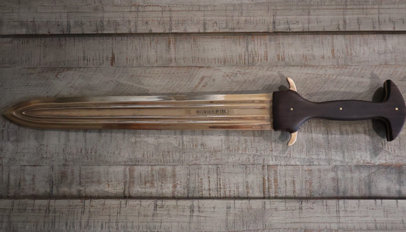 The finished sword in full: leaf-shaped bronze blade with parallel fullers, dark hardwood grip and flared pommel, laid across a weathered timber bench