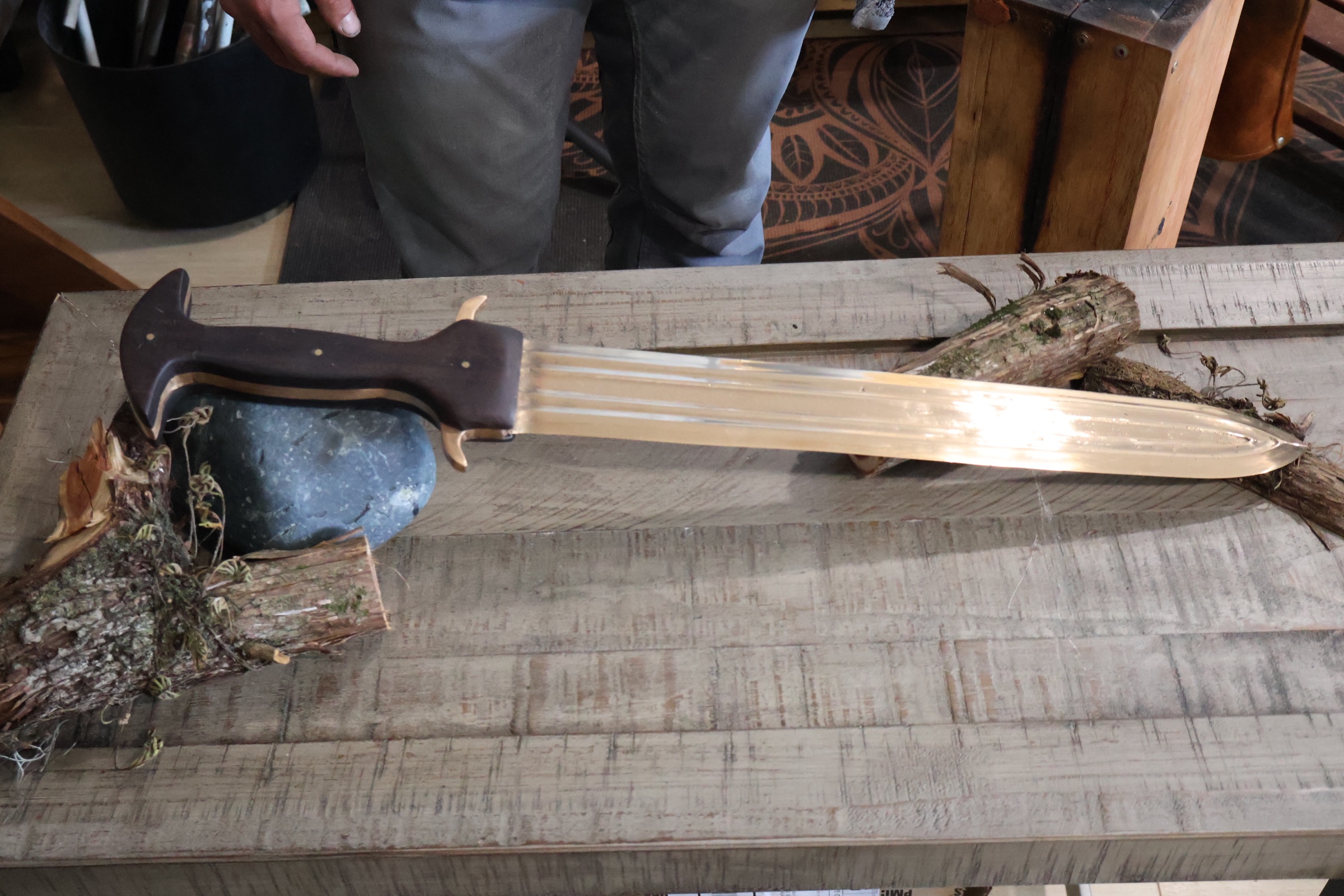The finished bronze sword laid on a timber bench beside a river stone and a length of mossy cut wood