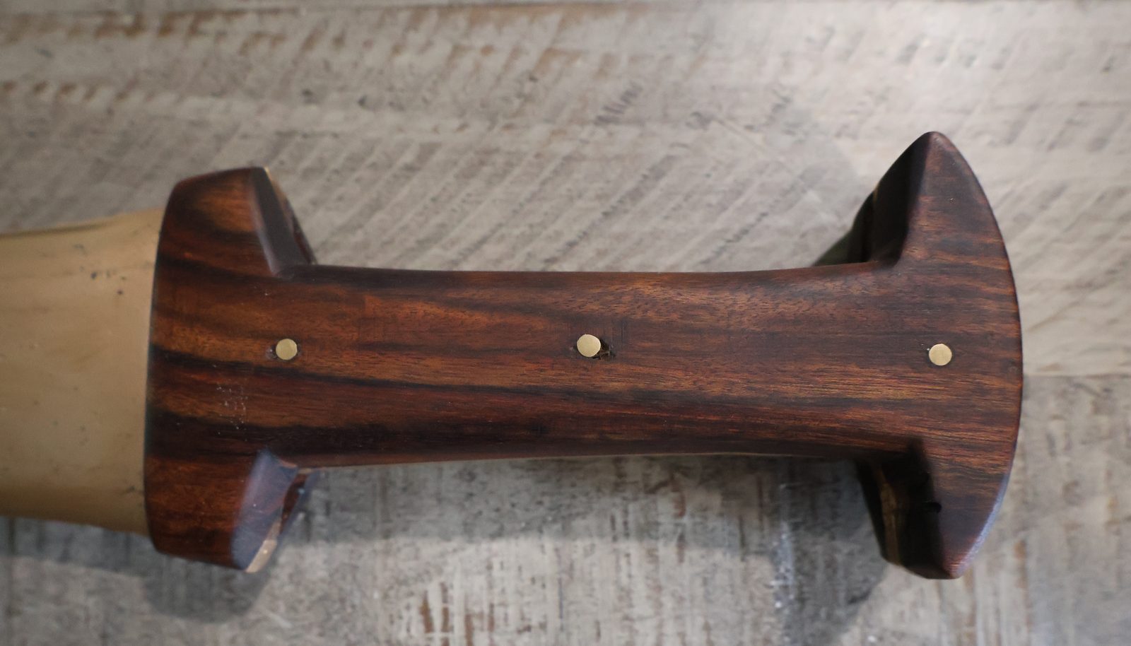 The grip seen from above: dark figured hardwood waisted at the middle, three brass pins in a line, flaring to a wide pommel