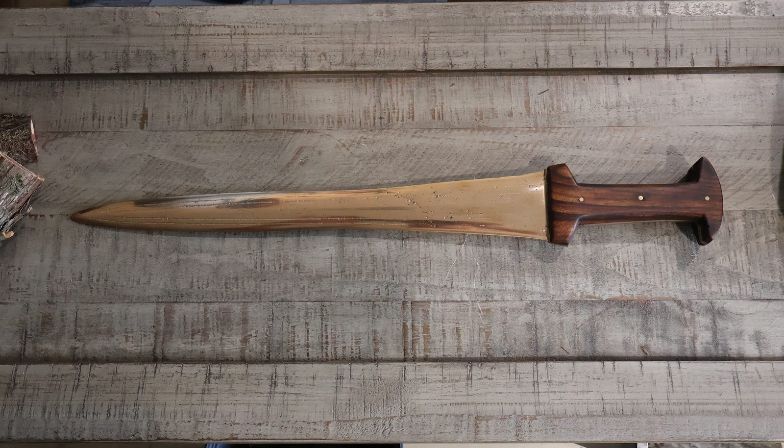 The finished sword in full on a timber bench, cut wood at one end and a river stone at the other